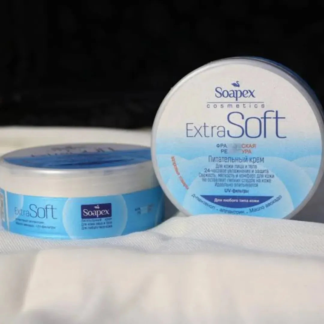 Two round tubs of Soapex Cosmetics ExtraSoft facial cream, one with a blue lid and one open showing the cream inside, set on a white textured surface against a dark background.