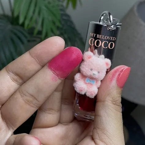A hand with pink nail polish holds a small lip gloss tube labeled 'MY DEARLY FAVORITE COCO' next to a pink plush bear charm. The background shows a blurred indoor setting with green plants.