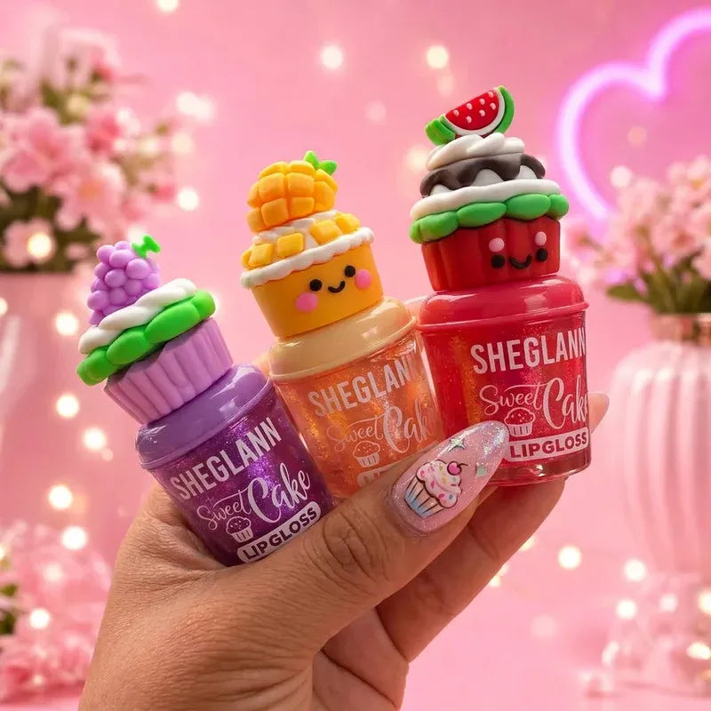 Three small lip gloss bottles shaped like cupcakes are held in a hand against a pink bokeh background. The bottles are labeled SHEGLANN Sweet Cake Lipgloss and come in purple, orange, and red colors with cute frosting and fruit toppings.