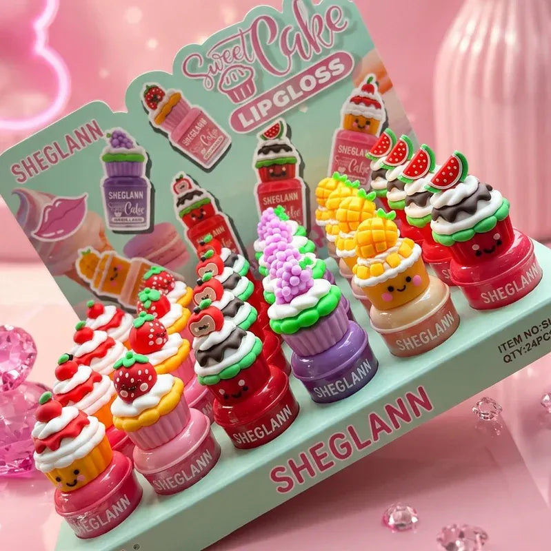 A display of cute, cupcake-shaped lipglosses by SHEGLANN on a matching mint green stand. The lipglosses are shaped like various colorful cupcakes with smiling faces, including strawberry, chocolate, vanilla, and watermelon flavors, set against a pink background with decorative pink elements.